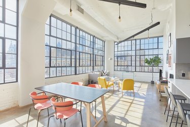 Modern loft living space with dining table, orange dining chairs, yellow accent chairs, gray couch, large industrial windows, concrete floors.